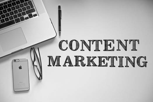 Černobílá fotka notebooku, mobilu, brýlí a pera na stole s nápisem Content Marketing, ilustrující obsahový marketing.