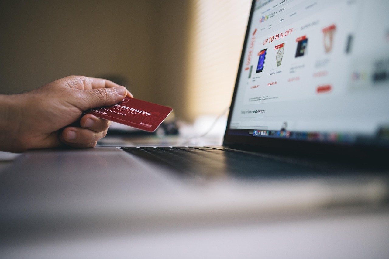 Ruka držící červenou platební kartu před notebookem s otevřeným e-shopem a slevou až 70 % při online nákupu